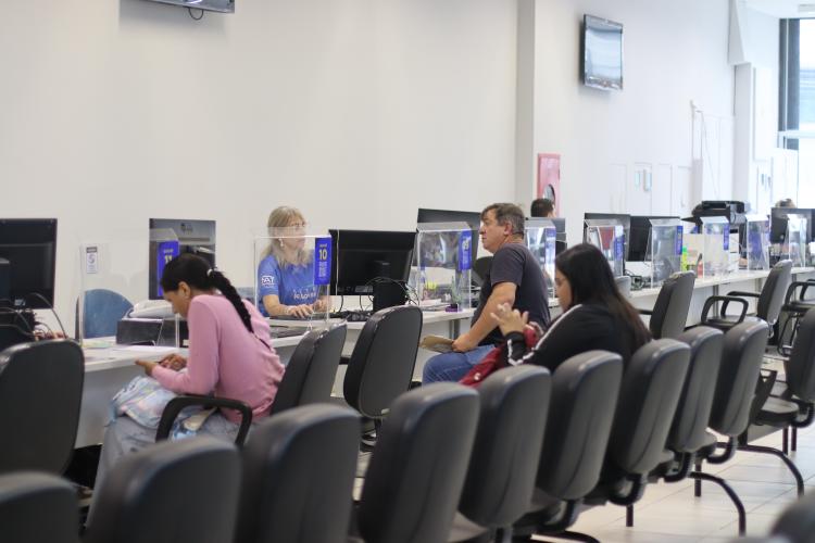 Ambiente interno da Casa do Trabalhador de Curitiba. Em primeiro plano, pessoas aguardam nas cadeiras de espera. Ao fundo, cidadãos recebem atendimento presencial de funcionários nos guichês com divisórias transparentes.