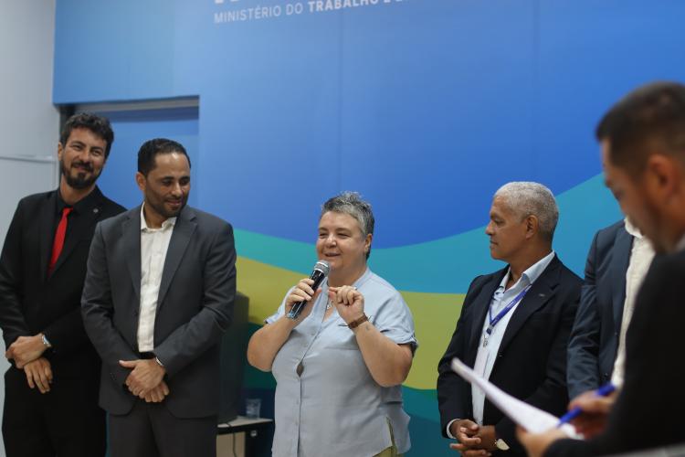 mulher falando ao microfone, enquanto outras pessoas a observam