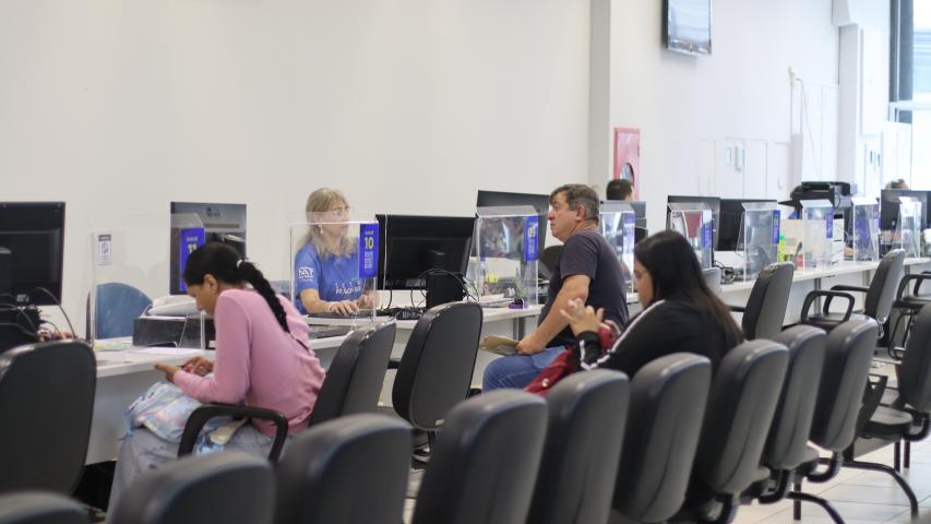 Ambiente interno da Casa do Trabalhador de Curitiba. Em primeiro plano, pessoas aguardam nas cadeiras de espera. Ao fundo, cidadãos recebem atendimento presencial de funcionários nos guichês com divisórias transparentes.